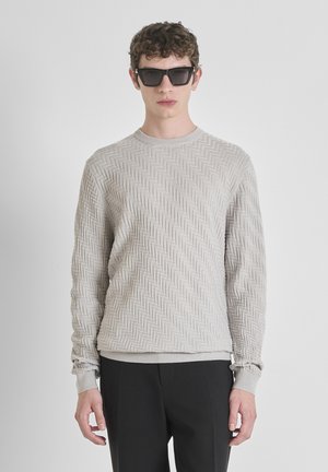 Maglione grigio chiaro con un motivo a chevron testurizzato, scollo rotondo e polsini a coste, abbinato a occhiali da sole neri, indossato con pantaloni neri.