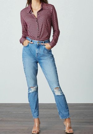 Rood geblokt overhemd met knopen en lange mouwen, gecombineerd met een high-waisted gespijkerde blauwe jeans en doorzichtige sandalen met hak.