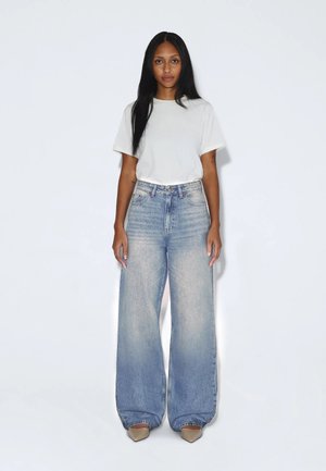 Vrouw met lang zwart haar, een wit T-shirt, lichtblauwe jeans met hoge taille en wijde pijpen, en beige puntige schoenen, staand tegen een effen achtergrond.