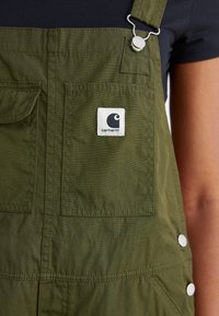 Olive green overalls made of durable fabric, featuring adjustable straps, a front pocket with a logo patch, and silver snap fasteners.