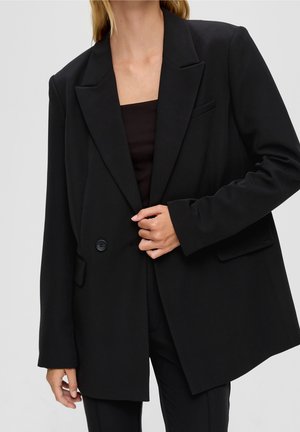 Frau trägt einen schwarzen oversized Blazer über einem schwarzen Top, knöpft einen Knopf, vor einfachem hellem Hintergrund.