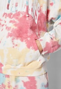 Personne portant un sweat à capuche tie-dye et un pantalon de survêtement aux couleurs rose, jaune et bleu, main dans la poche avant, texte partiel visible sur la poitrine.