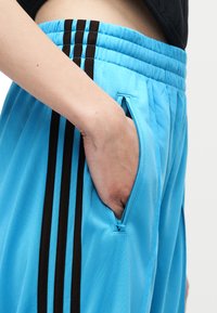 Χέρι στην τσέπη μπλε αθλητικών παντελονιών με μαύρες ρίγες και φερμουάρ στην πλαϊνή τσέπη, συνδυασμένα με μαύρο τοπ.