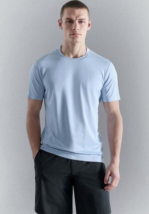 Giovane uomo con capelli corti che indossa una camicia celeste a maniche corte e pantaloncini neri, in piedi davanti a uno sfondo grigio uniforme.