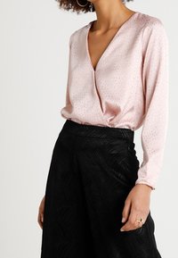 Blouse en satin rose clair avec un col en V et un motif à pois texturé, associée à une jupe plissée noire avec un design ondulé.
