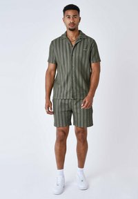 Camisetta verde a righe con maniche corte e colletto a scollo, abbinata a pantaloni corti coordinati. Indossata con sneakers bianche e accessori.