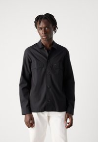 Camisa negra de manga larga confeccionada en tela suave, con cierre de botones en el frente, dos bolsillos en el pecho y cuello con solapas.