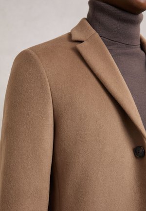 Mann trägt einen braunen Wollmantel über einem dunkelbraunen Rollkragenpullover, Fokus auf den Schulter- und Kragenbereich mit einem sichtbaren Knopf.