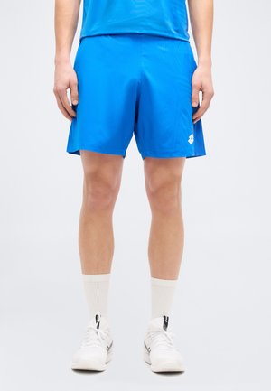 Férfi élénk kék sportnadrágban, fehér zokniban és fehér sportcipőben, aki egy egyszerű világos háttér előtt áll.