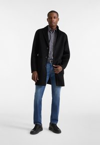 Abrigo de lana negro con cuello muesca, camisa a cuadros gris y rojo, jeans azules y zapatillas negras. Cuenta con botones y bolsillos frontales.