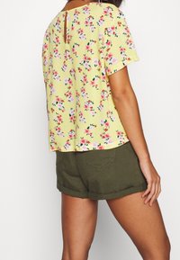 Hedvábná blůza s květinovým potiskem v žluté barvě, krátké rukávy, zadní otvor ve tvaru klíče a volnější střih, v kombinaci s olivově zelenými šortkami.