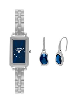 Silberne Metalluhr mit einem rechteckigen blauen Zifferblatt, silbernen Gliedern und weißen Stundenmarkierungen. Kombiniert mit blauen Anhängerohrringen in silbernen Fassungen.
