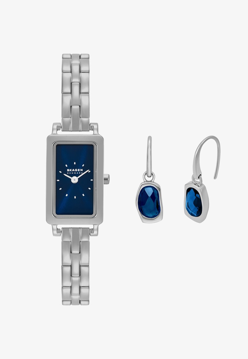 Reloj de metal plateado con una esfera rectangular azul, eslabones plateados y marcadores de hora blancos. Combinado con pendientes de gota de gema azul en monturas de plata.