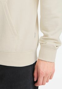 Felpa beige con tasca frontale, polsini a costine e una piccola etichetta sul lato. Il materiale ha una texture morbida.