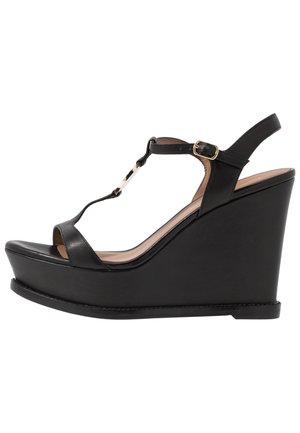 Sandalia de cuña de cuero negro con tira en T, hebilla dorada, punta abierta y tacón alto sobre fondo blanco.