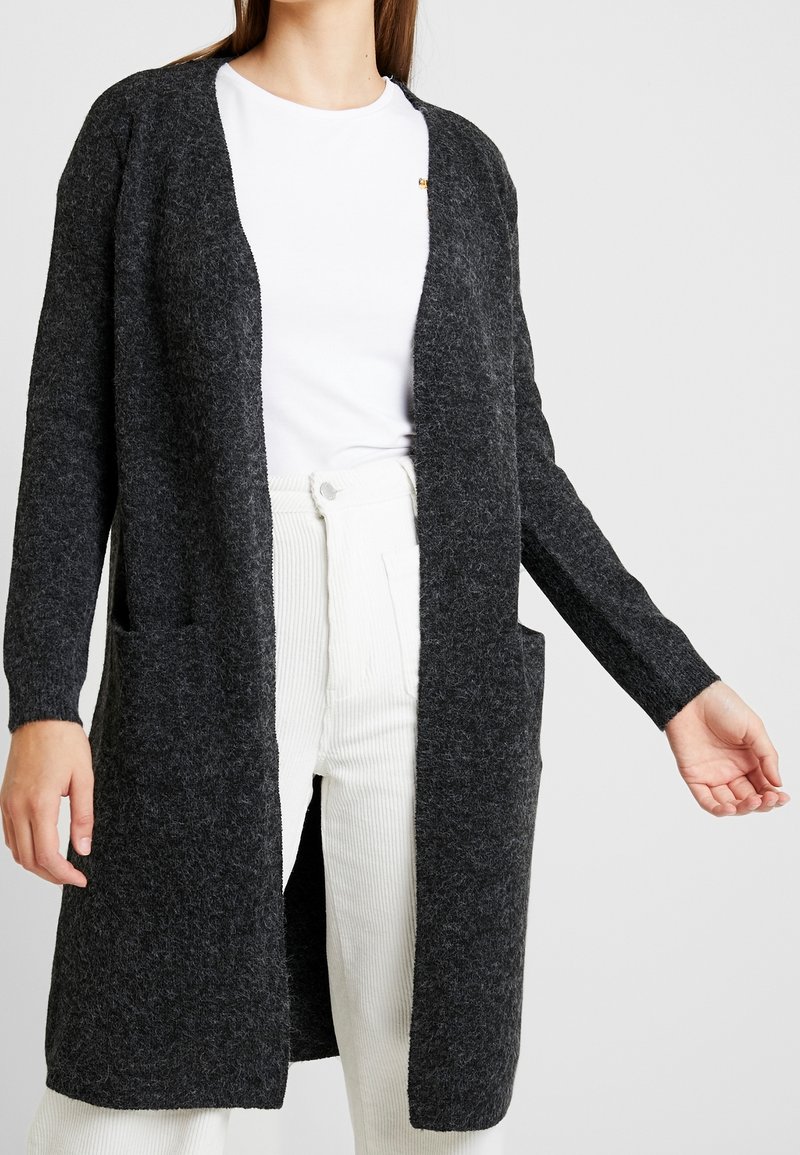 Femme portant un long cardigan gris foncé ouvert avec des poches sur un t-shirt blanc et un pantalon blanc, sur un fond clair uni.