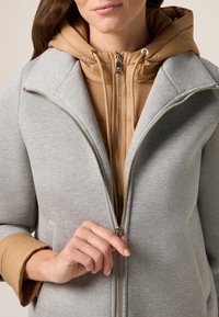 Cappotto grigio con zip e strato interno a cappuccio color sabbia; presenta un colletto prominente, una texture liscia e dettagli in zip argentati.