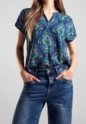 Nainen, jolla on sininen kuvioitu lyhythihainen pusero, joka on tönäisty sinisiin farkkuihin, seisoo käsivarret taskuissa yksiväristä taustaa vasten.