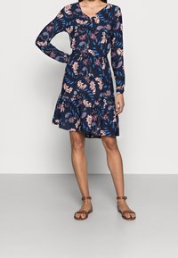 Robe florale navy à manches longues, décolleté en V, taille élastique et jupe à volants. Présente des motifs floraux roses et bleus. Portée avec des sandales marron.