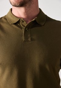 Polo de color verde oliva hecho de tejido texturizado, con un cuello clásico y una tapeta de tres botones. Mangas cortas.