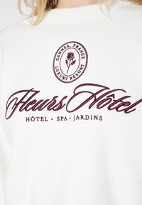 Felpa bianca con ricamo bordeaux che riporta "Cannes, France Luxury Resort" e "Fleurs Hôtel SPA Jardins." Tessuto morbido, design con colletto a girocollo.