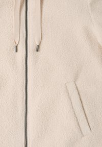 Giacca in pile beige con trama, chiusura frontale con zip, due cordini con punte metalliche e tasca laterale inclinata a destra.