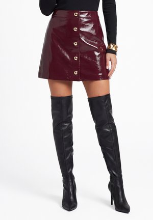 Donna che indossa una mini gonna bordeaux lucida con bottoni sul davanti, top nero a maniche lunghe, bracciale dorato e stivali neri con tacco sopra il ginocchio.