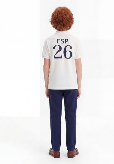 Παιδί με σγουρά κόκκινα μαλλιά στέκεται με την πλάτη προς τα εμπρός, φοράει λευκό πόλο με το «ESP 26» και σκούρο μπλε παντελόνια και καφέ παπούτσια.