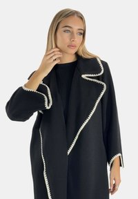Black coat with a textured finish, featuring a white scalloped trim, wide lapels, and loose sleeves. The design is long and open-fronted.