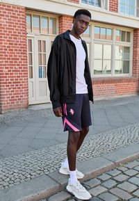 Fekete dzseki, fehér póló, sötét rövidnadrág rózsaszín részletekkel és világos edzőcipő. Texturált anyag és sportos dizájn jellemzi. Kültéri környezet.