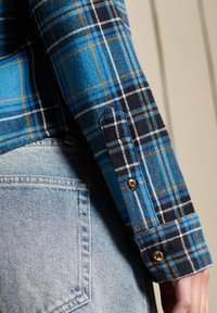 Camicia in flanella con un motivo a quadri blu, nero e arancione. Presenta polsini con bottoni e una texture morbida, abbinata a denim blu chiaro.