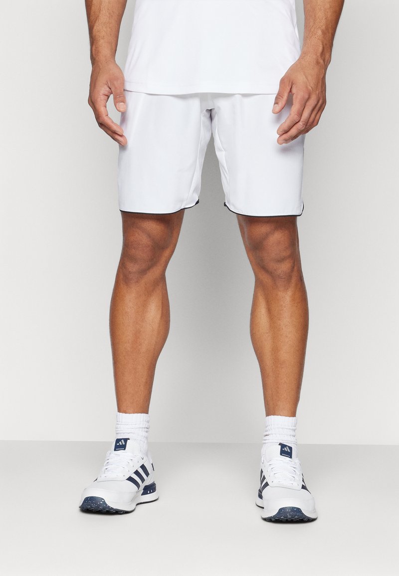 Pantaloni sportivi bianchi con bordi neri, abbinati a scarpe sportive bianche e blu navy. Modello mostrato in piedi, braccia rilassate lungo i fianchi.
