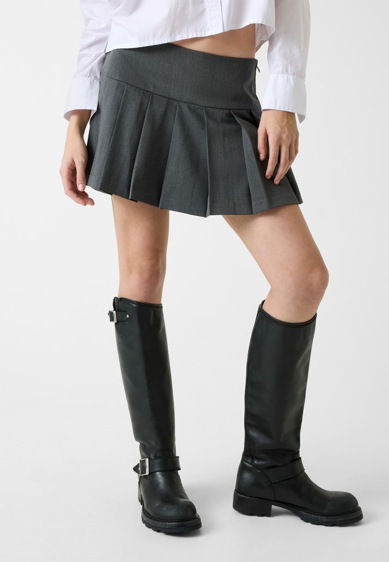 Mini-jupe plissée grise avec une ceinture haute, associée à de grandes bottes en cuir noir avec des boucles et une surface texturée.