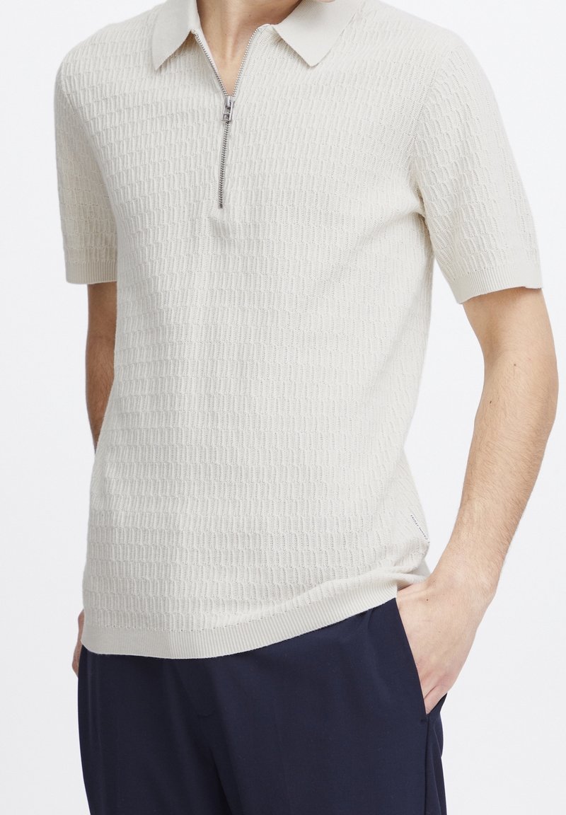 Homme portant un polo en maille texturée beige clair à manches courtes avec une fermeture éclair argentée à l'avant et un pantalon bleu marine, main dans la poche.