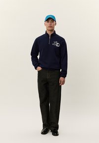 Jovem em pé, com postura ereta, vestindo um sweatshirt azul-marinho com fecho de meio zíper, calças pretas, boné azul e sapatos pretos, contra um fundo simples.