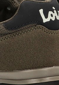 Zapatilla de ante marrón con acentos negros, superficie texturizada, detalles de costura y suela plana gris. Logotipo impreso en el costado.