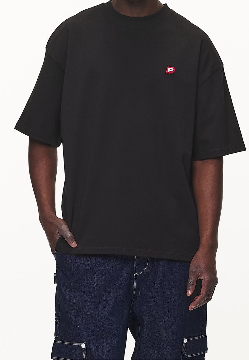 Camiseta negra de gran tamaño fabricada en algodón, con mangas cortas y un logo rojo bordado en el pecho izquierdo. Diseño sencillo y ajuste relajado.