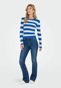 Haut rayé bleu et blanc à manches longues avec un tissu côtelé, associé à un jean foncé évasé et des baskets blanches, se tenant contre un fond neutre.