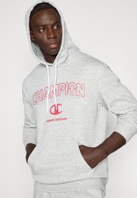 Γκρι φούτερ με κουκούλα και κόκκινο λογότυπο με το κείμενο "CHAMPION" και μικρότερη επιγραφή "Authentic Athleticwear." Διαθέτει μπροστινές τσέπες και κουκούλα με κορδόνια.