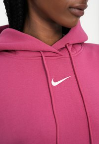 Roza kapuca iz mehke tkanine, ki ima bel Nike logotip, vrvico in prostorno kapuco. Podroben posnetek na predelu vratu in ramen.