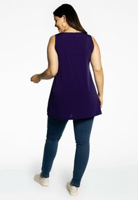 Lilla ærmeløs top med en afslappet pasform, der har en rund halsudskæring og en længere bagkant, parret med mørkeblå skinny jeans og beige sneakers.