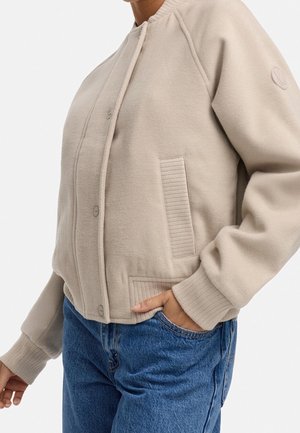 Henkilö pukeutunut beigen väriseen napitettuun bomber-takkiin, jossa on resorit hihansuissa ja taskujen yksityiskohdat, yhdistetty sinisiin farkkuihin, käsi taskussa.