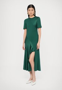 Smaragdgroene, knielange jurk met korte mouwen; voorzien van diagonale accenten en een voorsplit. Gladde, lichte stof; gecombineerd met witte hakken.