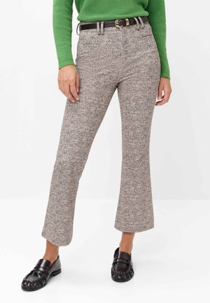 Frau trägt beige karierte kurze Hose, schwarzen Gürtel, grünen Pullover und schwarze Loafers und steht vor weißem Hintergrund.