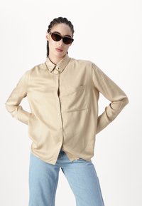 Ljusbeige blus i ett mjukt tyg, knäppning framtill, krage och en bröstficka. Kombineras med ljusblå jeans och svarta solglasögon.