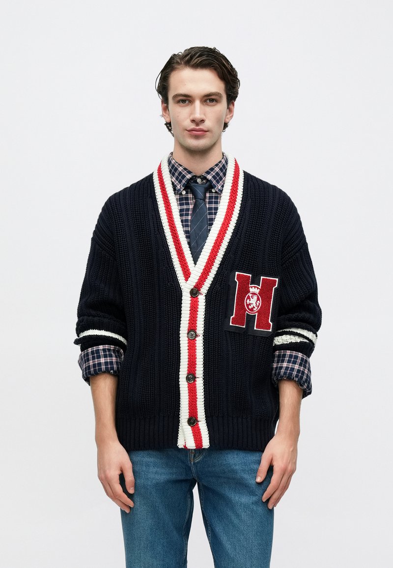 Μπλε ναυτικό πλεκτό ζακέτα με λευκές και κόκκινες ρίγες, διακοσμημένη με ένα μεγάλο κεντημένο έμβλημα 'H', φορεμένη πάνω από ένα καρό πουκάμισο και γραβάτα.