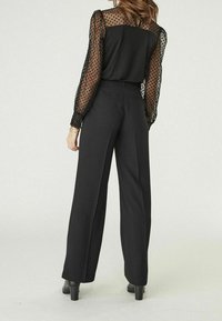 Blouse noire avec manches transparentes à pois, associée à un pantalon noir taille haute à jambes larges et des bottes à talons, vue de dos.