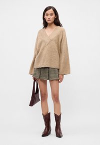 Mulher vestindo um suéter em creme felpudo de decote V, saia mini plissada xadrez, segurando uma bolsa bordô e botas de salto midi bordô, em pé sobre fundo branco.