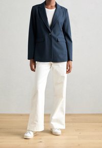 Námořnický blazer s jedním knoflíkem, dvěma předními kapsami, spárovaný s bílými zvonovými kalhotami a bílými teniskami na dřevěné podlaze.