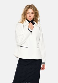 Weißer Pullover-Jacke mit Kapuze, halbem Reißverschluss auf der Vorderseite und schwarzen Streifenakzenten an den Ärmelbündchen und Taschen. Glatte Textur, lässiges Design.